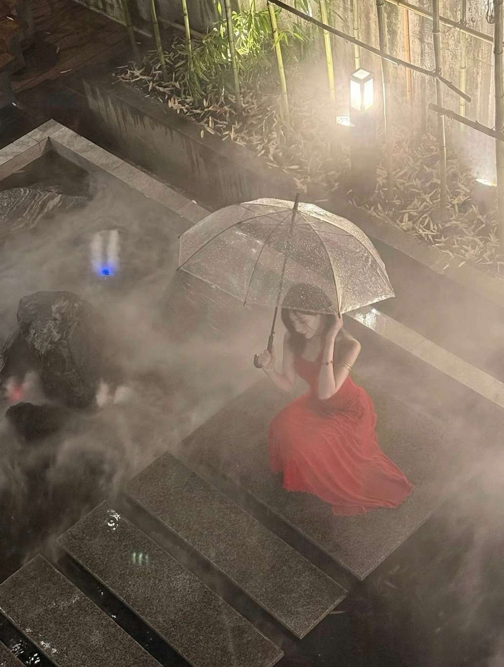 Maram in a red dress crouching on stone steps with a clear umbrella in mist