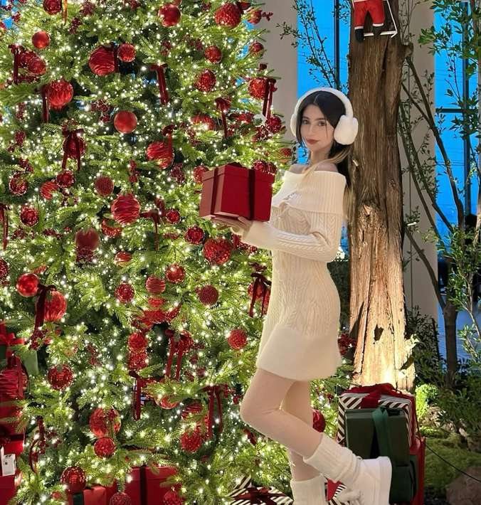Maram in a cream knit dress holding a red gift by a Christmas tree