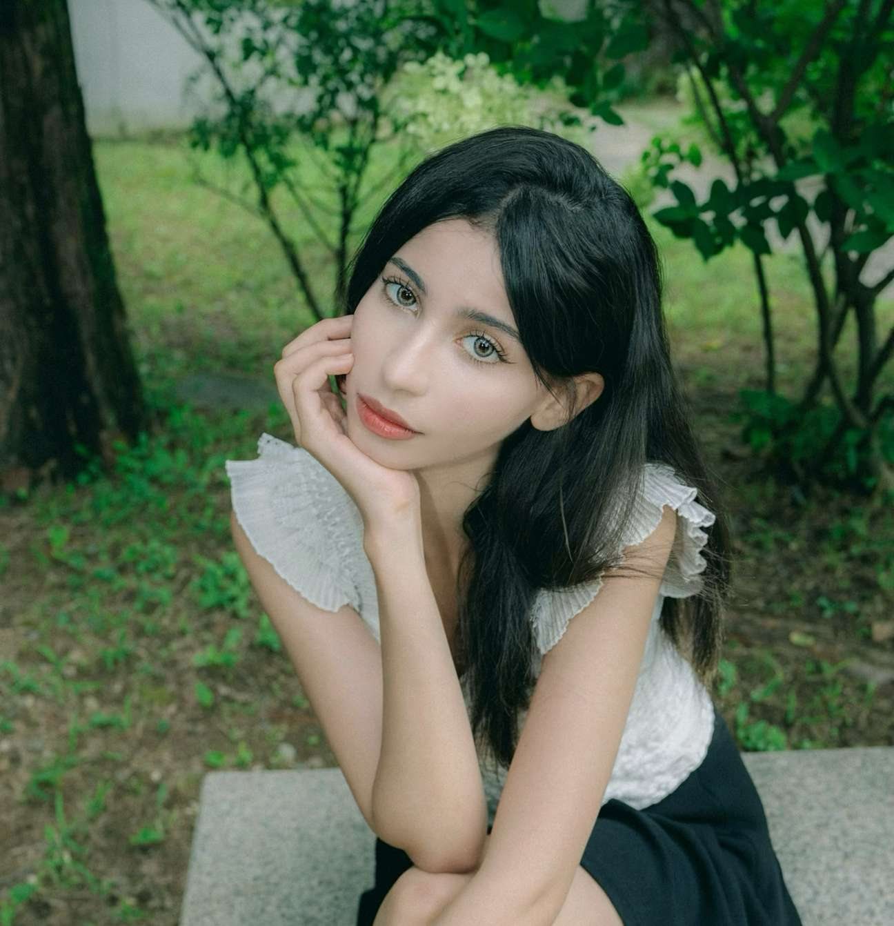 Maram resting her chin on her hand, seated outdoors in a white ruffled top